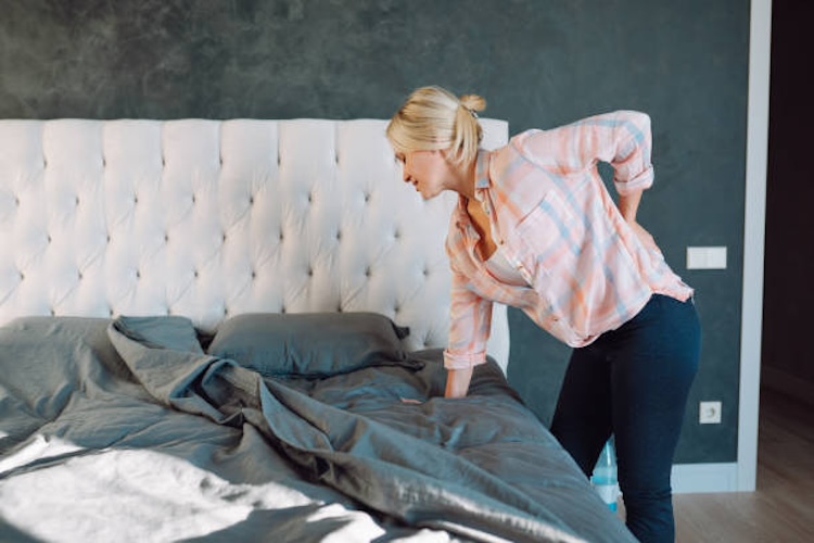 Blond woman holding her back and incline on bed in bedroom. Side view